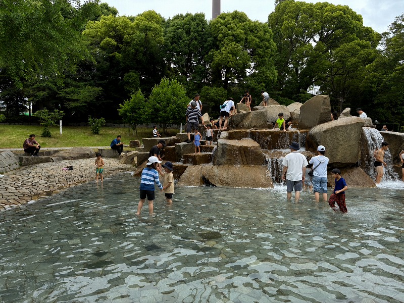 平塚市総合公園「流れの広場」の岩造りの滝。浅い水辺で家族連れが水遊びを楽しんでいる様子